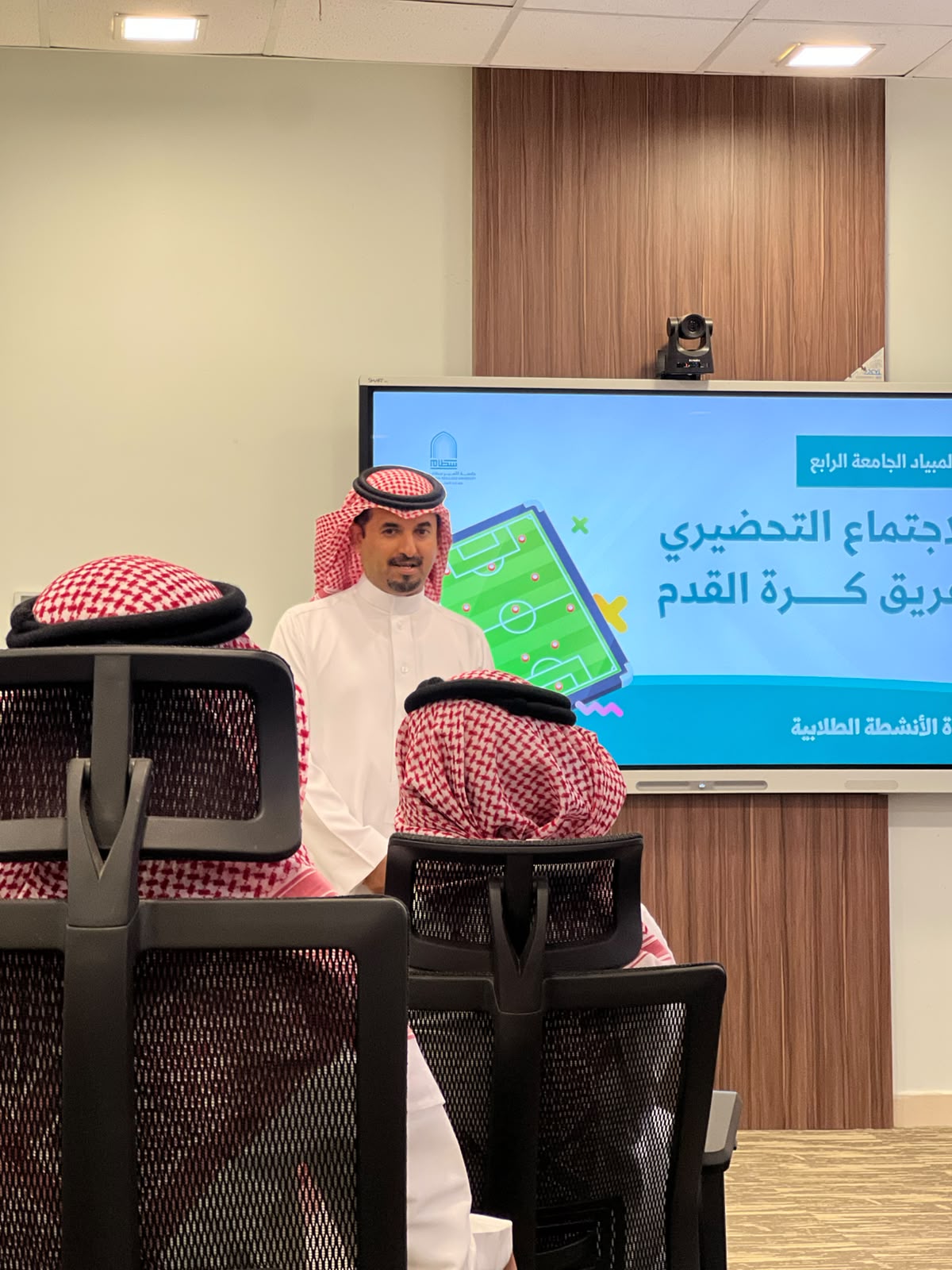 متحدث يخاطب الطلاب أمام شاشة الاجتماع التحضيري لفريق كرة القدم ضمن الأولمبياد الجامعي الرابع