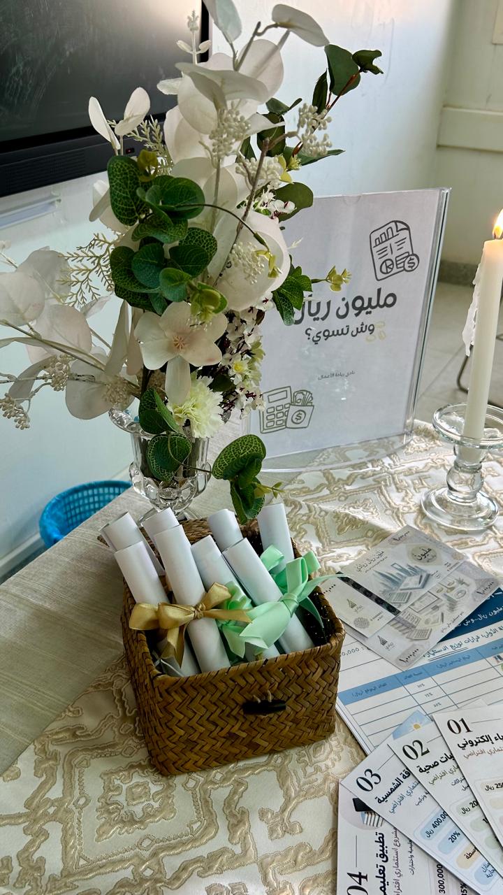 طاولة ركن فعالية "مليون ريال وش تسوي؟" تضم لوحة تعريفية وسلة أوراق ملفوفة ونشرات استثمارية